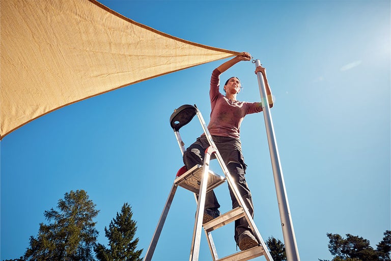 En kvinna monterar ett solsegel på en stege på en stolpe.