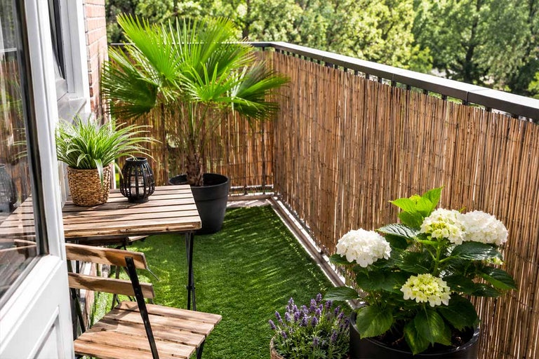 Organiserad, ljus balkong med insynsskydd av bambu, konstgräs, bistroset i trä och olika krukväxter som en palm och hortensia.