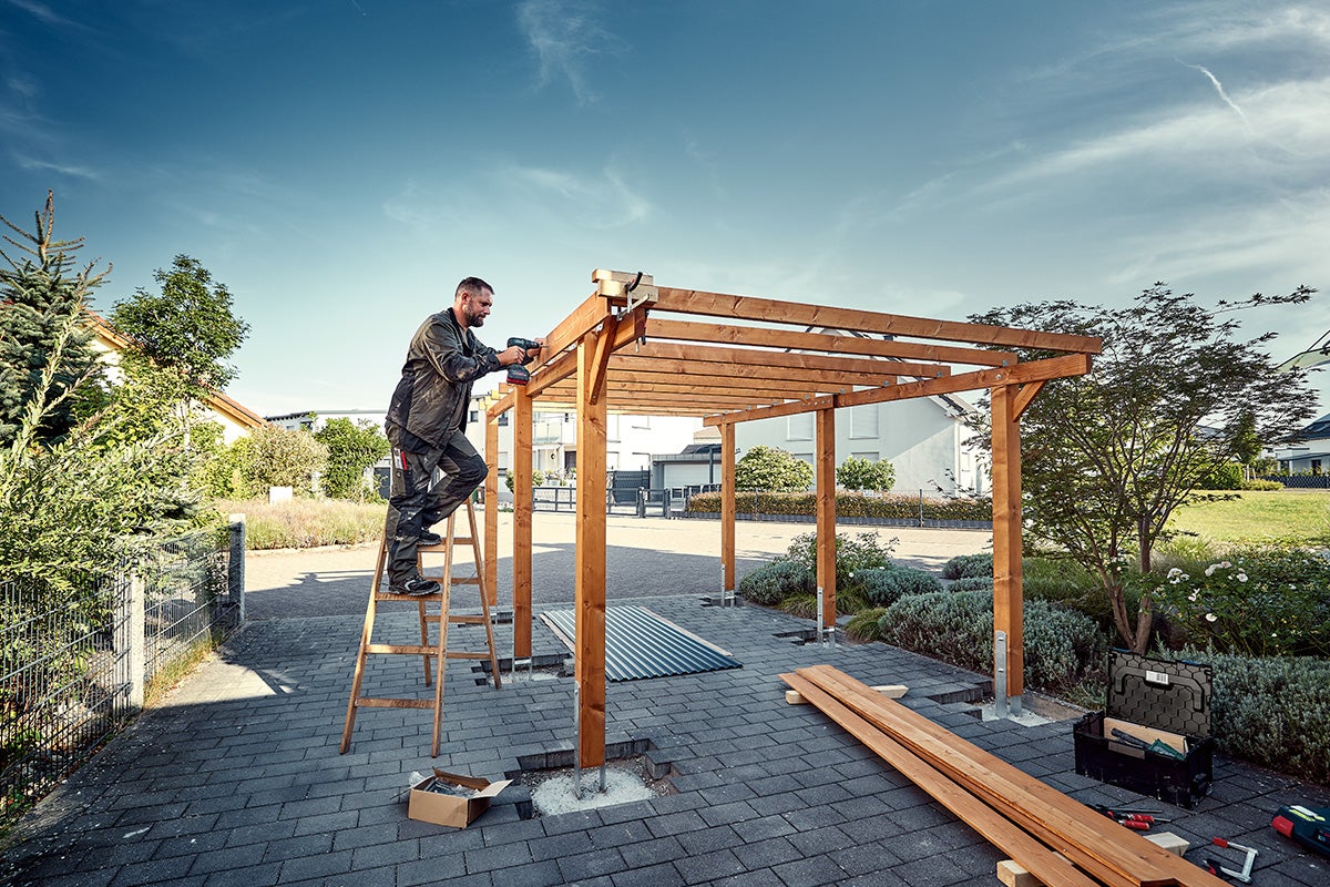 En man monterar en träpergola i trädgården med en batteridriven skruvdragare, stående på en stege.