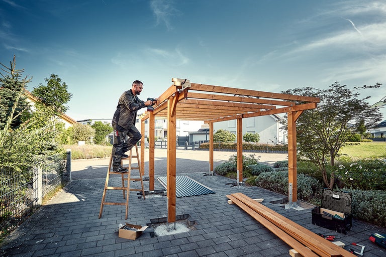 En man monterar en träpergola i trädgården med en batteridriven skruvdragare, stående på en stege.