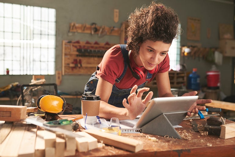 Kvinna med surfplatta i en verkstad, omgiven av verktyg och material för träbearbetning.