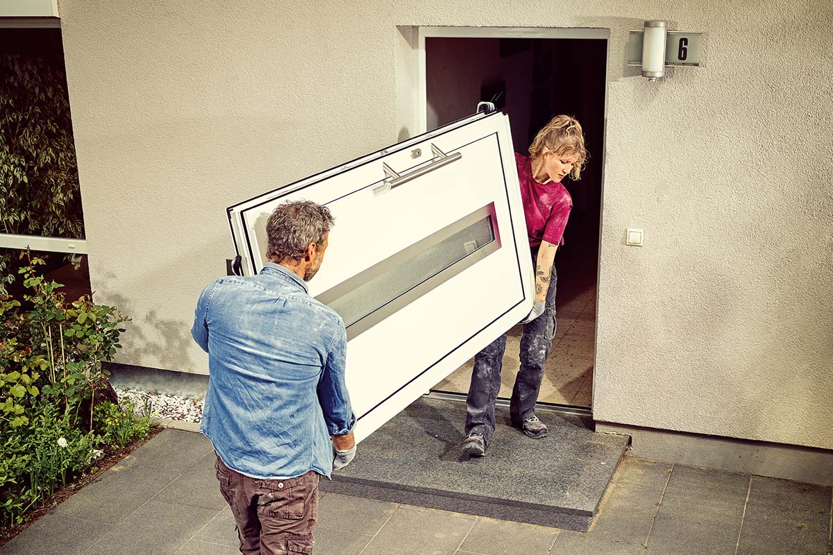 Två personer bär en ytterdörr med glasinsats till ett hus.