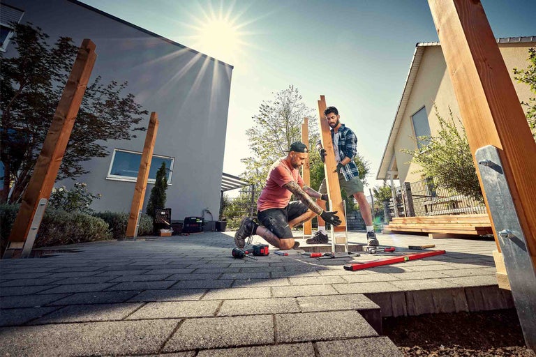 Två hantverkare bygger en träkonstruktion i trädgården med verktyg som en borrmaskin, vattenpass och skruvtvingar.