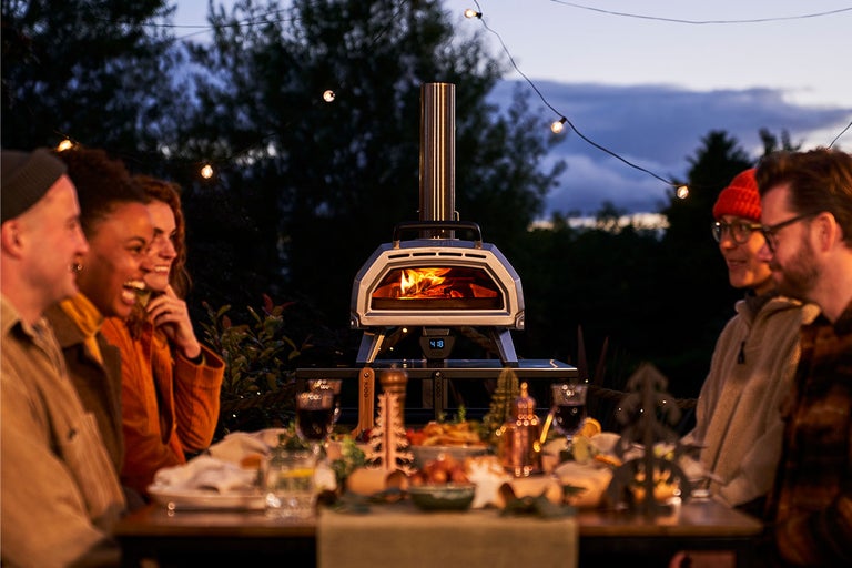 Grupp av människor sitter runt ett bord med en pizzaugn utomhus på natten
