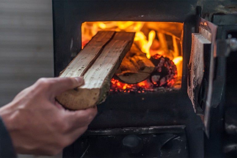 En hand lägger ved i en vedeldad spis där det redan brinner en eld.