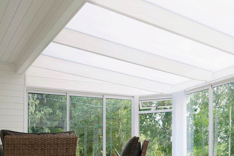 Ljust uterum med vita träbalkar, ljusgenomsläppliga takskivor och stora glaspartier med utsikt mot grönska.