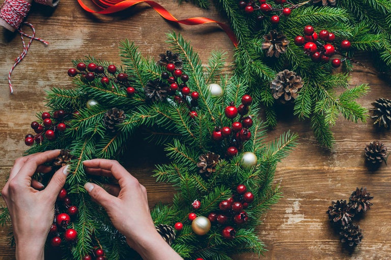 Dekorativ julkrans med grangrenar, bär och kottar arrangeras av händer på ett träbord.