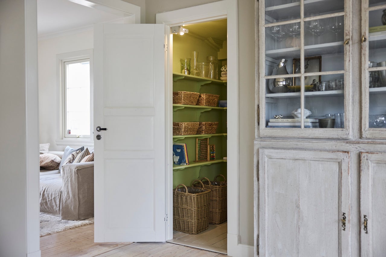 Ljust, organiserat skafferi med gröna trähyllor och flätade korgar, sett genom en vit dörr bredvid ett rustikt skåp.