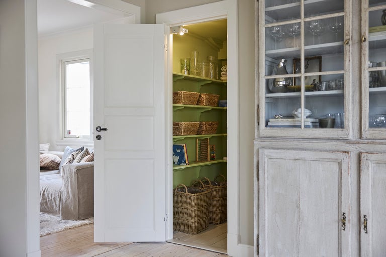 Ljust, organiserat skafferi med gröna trähyllor och flätade korgar, sett genom en vit dörr bredvid ett rustikt skåp.