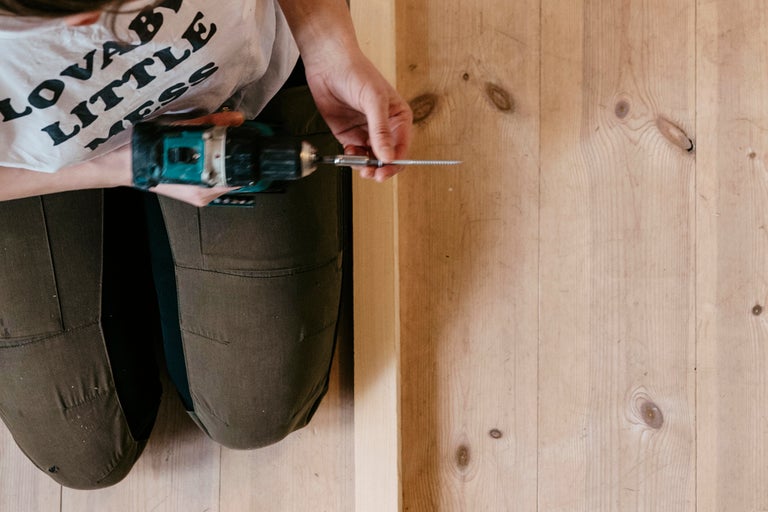 En person knäböjer med en batteridriven skruvdragare och en skruv framför en trävägg