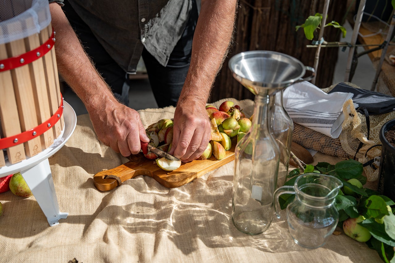 En man skär äpplen på en träskärbräda som förberedelse för en fruktpress med glasflaskor och tratt på ett bord.