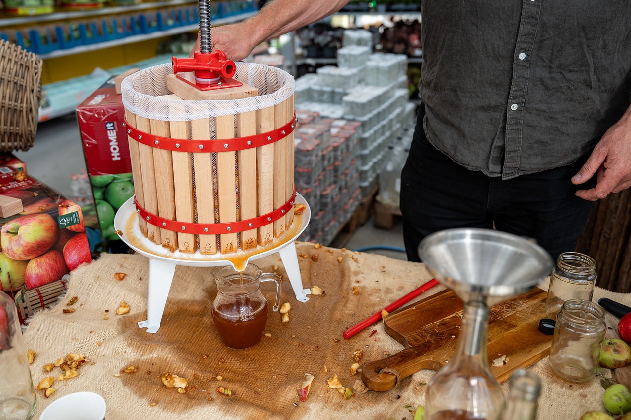 En fruktpress av trä med stålram som pressar ut färsk juice i en glaskaraff.