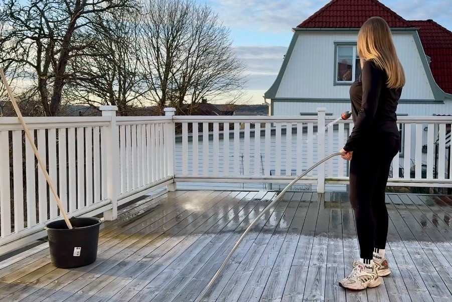 Rengöring av en träterrass med trädgårdsslang, sprutmunstycke, hink och skurborste på en terrass med vitt staket.