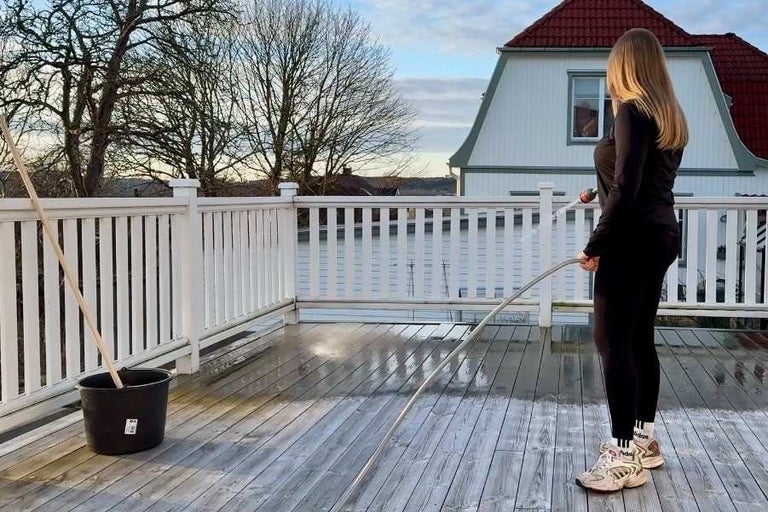Rengöring av en träterrass med trädgårdsslang, sprutmunstycke, hink och skurborste på en terrass med vitt staket.
