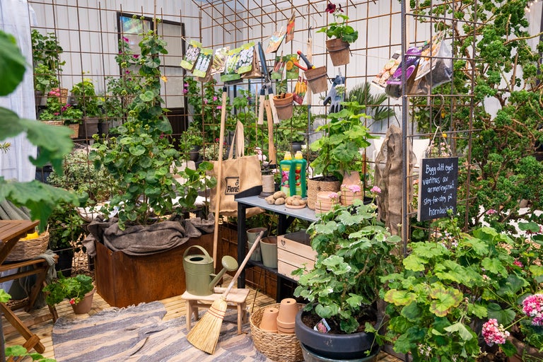 Ljus trädgårdshörna med växter i krukor, armeringsnät som spaljé, trädgårdsredskap, fröpåsar och väska med Flora Logo.