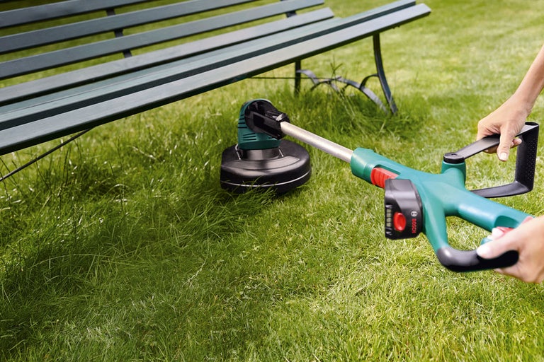 Person använder Bosch grästrimmer under en trädgårdsbänk på en gräsmatta.