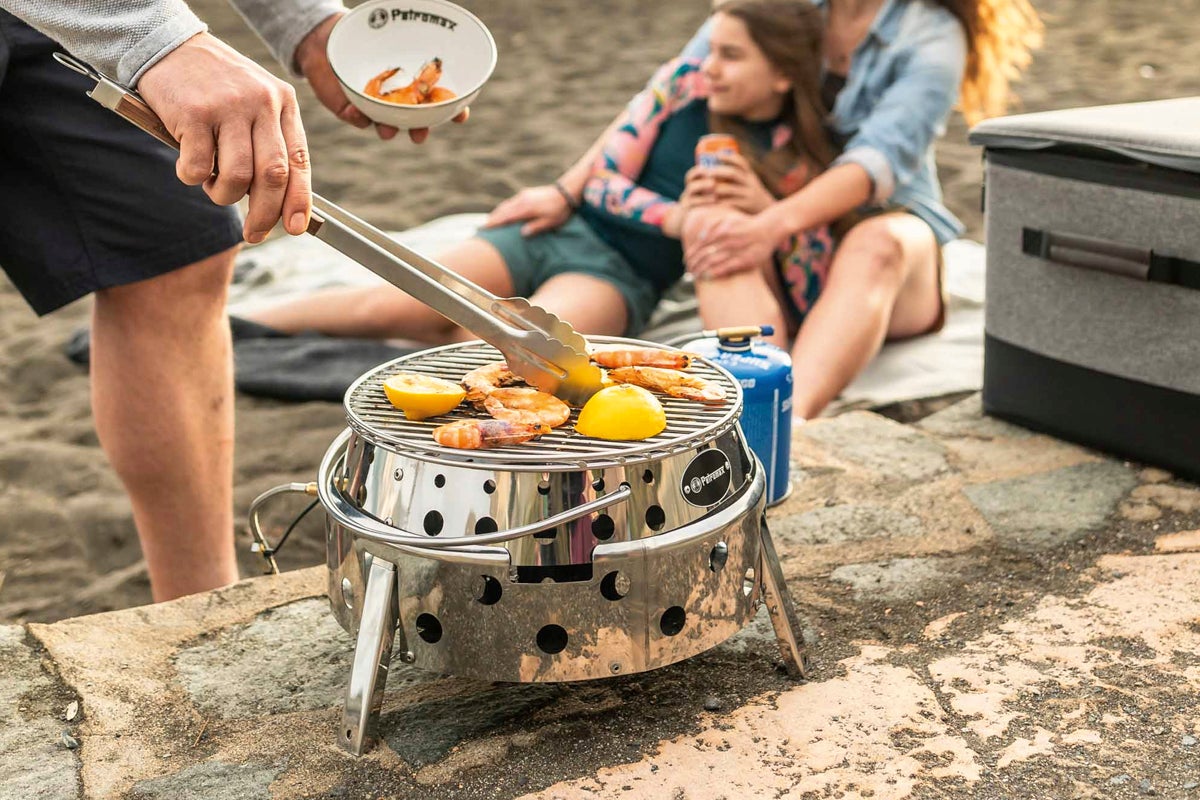Bärbar gasolgrill i rostfritt stål från Petromax i en ljus strandmiljö för grillning av räkor och citroner.