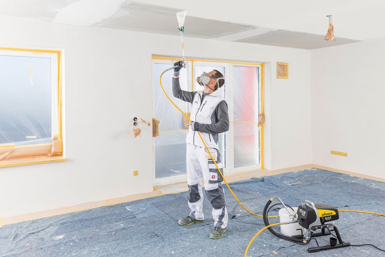 Person med andningsmask och färgspruta målar tak i ett förberett rum