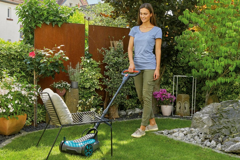 En kvinna använder en kompakt batteridriven gräsklippare under en trädgårdsstol på en grön gräsmatta i en trädgård.