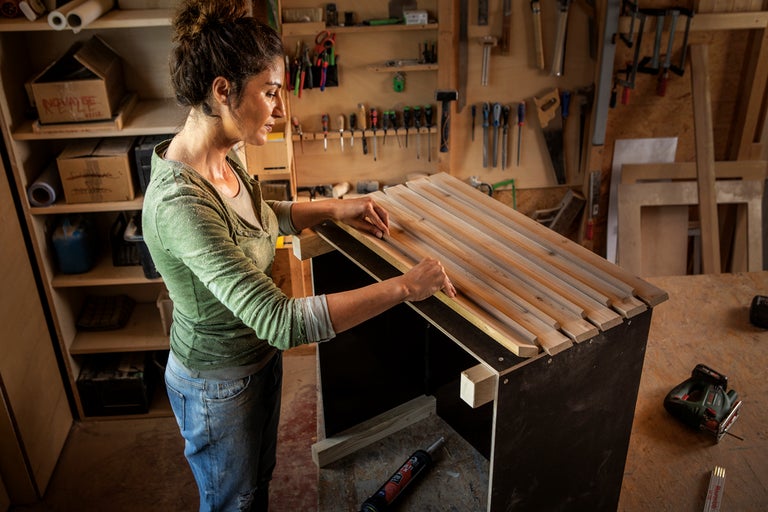 En kvinna monterar träribbor på en möbel i en verkstad med verktygsvägg, sticksåg och monteringslim.