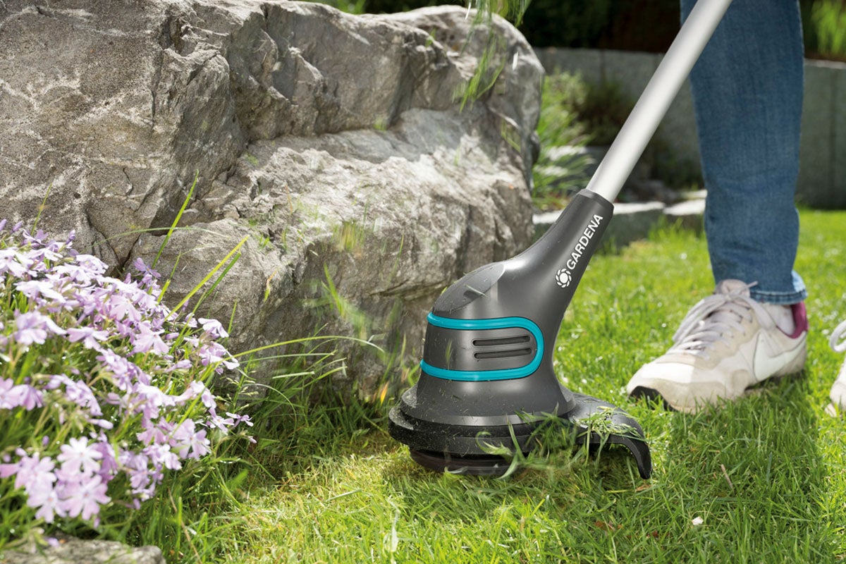 Gardena grästrimmer i bruk i en trädgård med blomsterrabatt och stenar