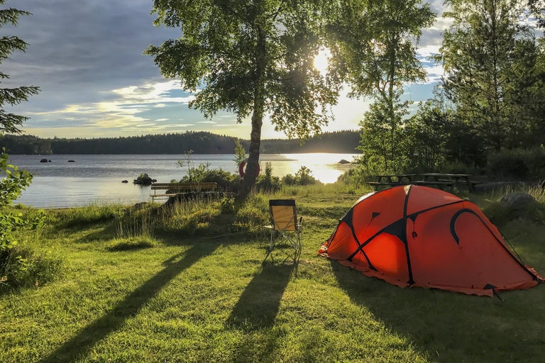 Scen med ett orange tält, en stol och ett träd vid en sjöstrand.