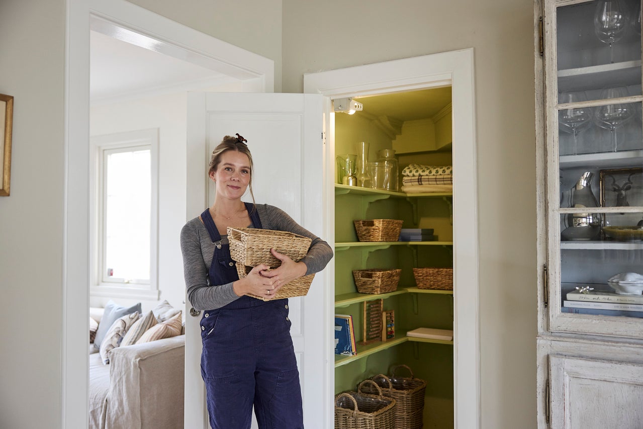 Kvinna håller en flätad korg framför ett upplyst skafferi med gröna hyllor och förvaringskorgar.
