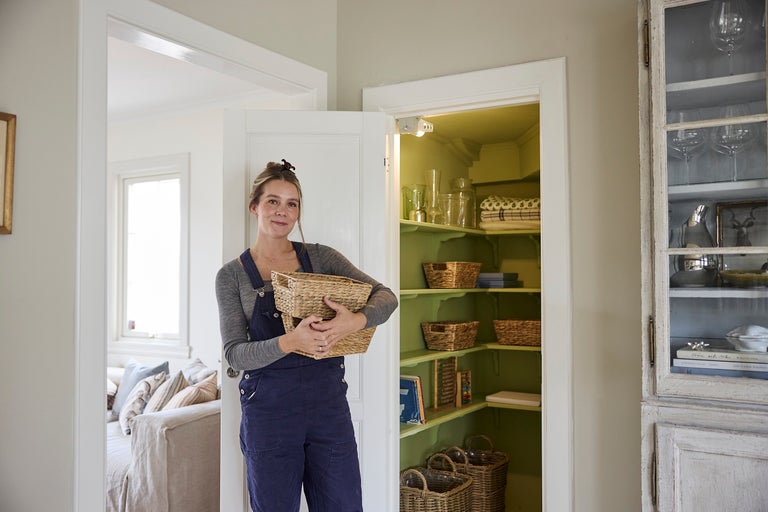 Kvinna håller en flätad korg framför ett upplyst skafferi med gröna hyllor och förvaringskorgar.