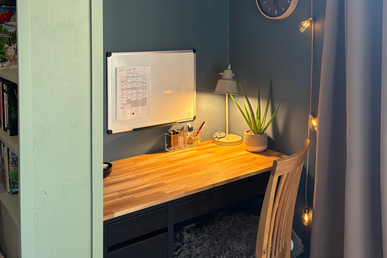 En organiserad arbetsplats med ett skrivbord i trä, skrivbordslampa, whiteboard och en Aloe Vera växt i en nisch.
