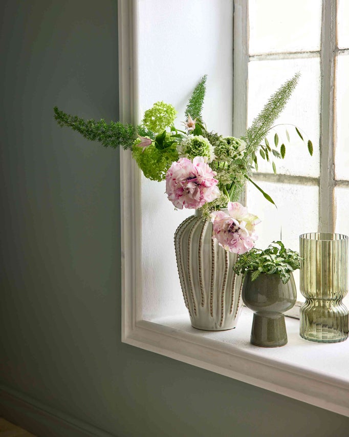 Blomvas med blommor, liten keramikruka med växt och en räfflad grön glasvas på en fönsterbräda.