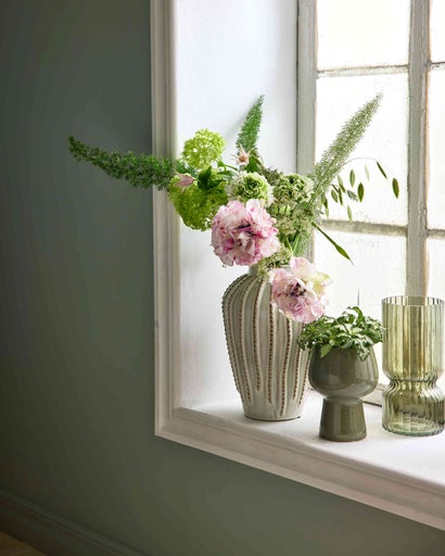 Blomvas med blommor, liten keramikruka med växt och en räfflad grön glasvas på en fönsterbräda.