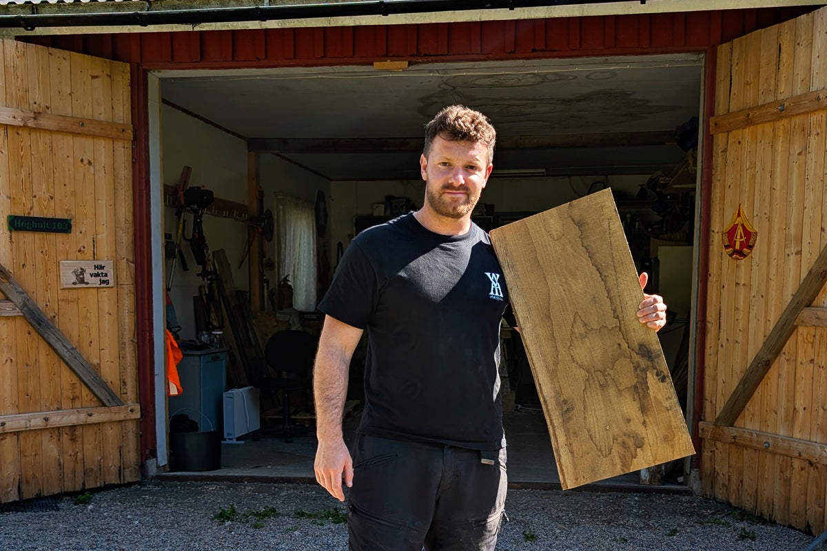 En man som håller en stor, betsad träskiva framför en öppen verkstad.