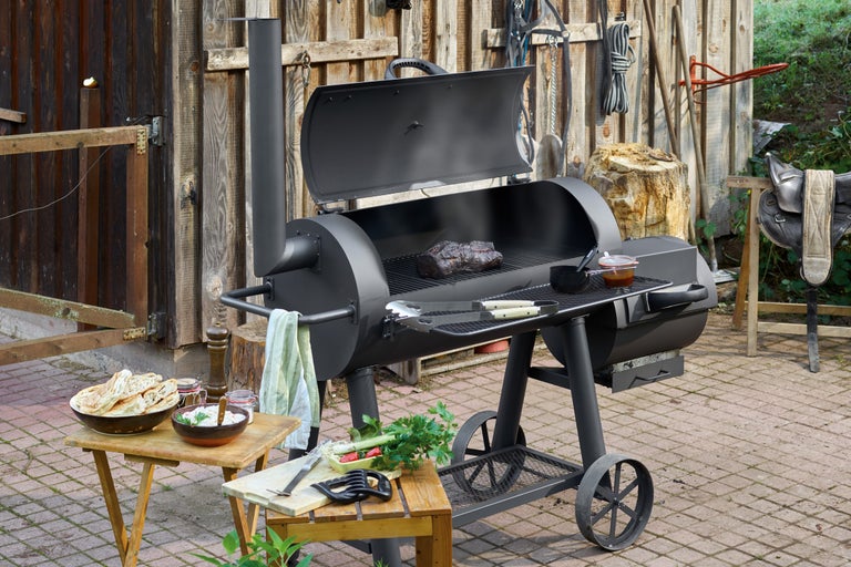 Svart rökgrill med mat och tillbehör på en gård.