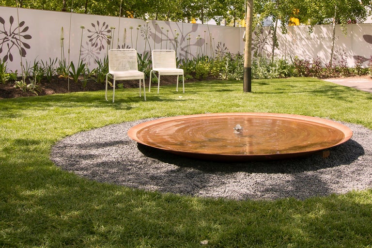 Trädgårdsscen med fontän, grusyta, gräsmatta, två stolar och planterat staket.