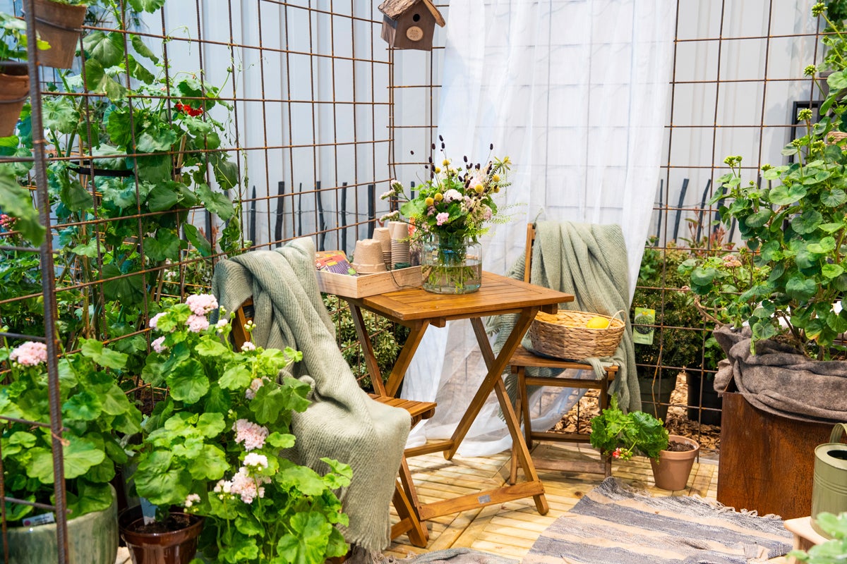 Ljus uteplats med fällbart träbord, stolar och nätstängsel i metall. Dekorerad med krukväxter, filtar, en vildblomsbukett och gardin.