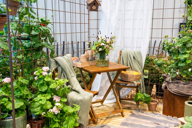 Ljus uteplats med fällbart träbord, stolar och nätstängsel i metall. Dekorerad med krukväxter, filtar, en vildblomsbukett och gardin.