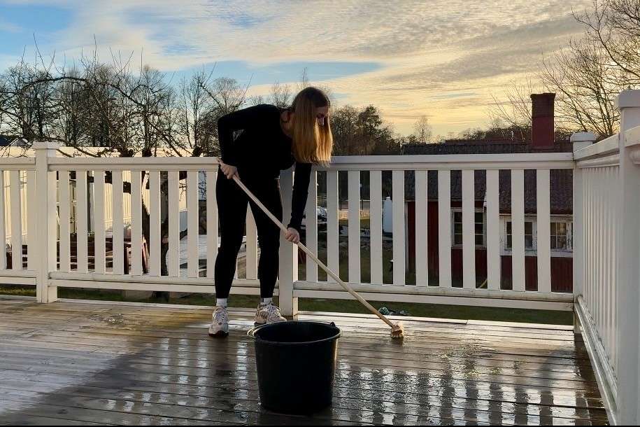En person rengör en träterrass med en skurborste och en hink. Terrassen har ett vitt träräcke.