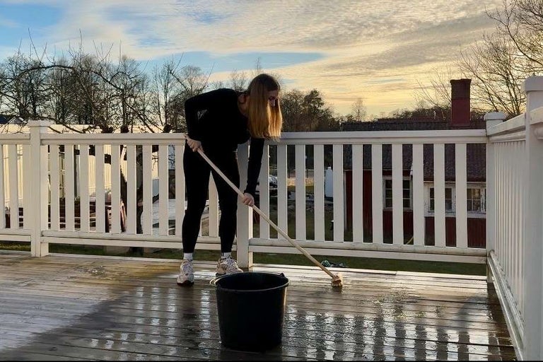 En person rengör en träterrass med en skurborste och en hink. Terrassen har ett vitt träräcke.