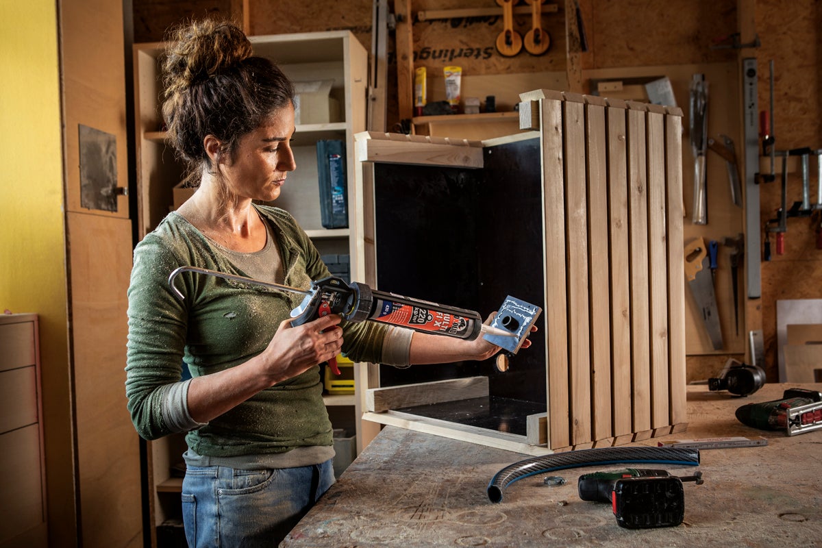 En kvinna applicerar monteringslim med en patronspruta på en del till en trälåda i en verkstad.