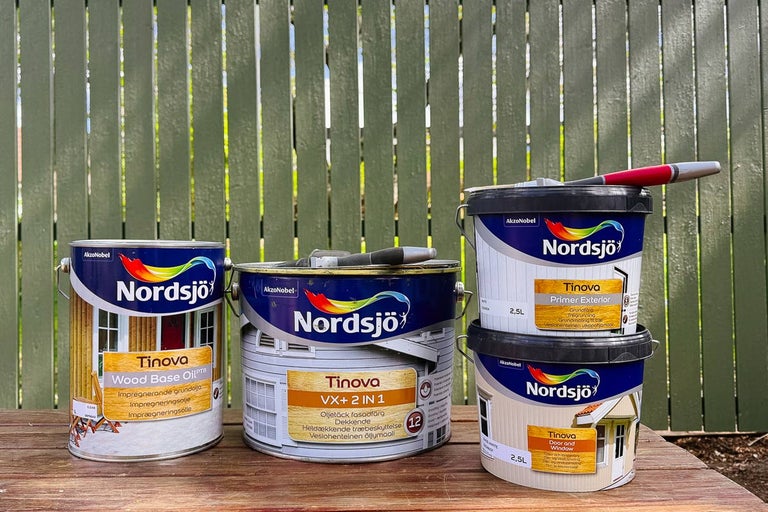 Flera burkar Nordsjö Tinova träfärg för utomhusbruk på ett träbord framför ett grönt trästaket