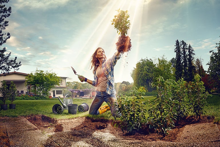 En kvinna knäböjer i trädgården och håller en växt med rotklump i handen för att plantera den.