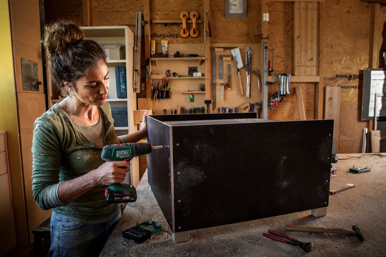 En kvinna monterar en svart trälåda med en skruvdragare i en verkstad fylld med verktyg.
