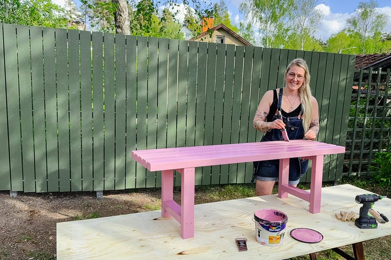 En kvinna målar en träbänk med pensel och färg i trädgården.
