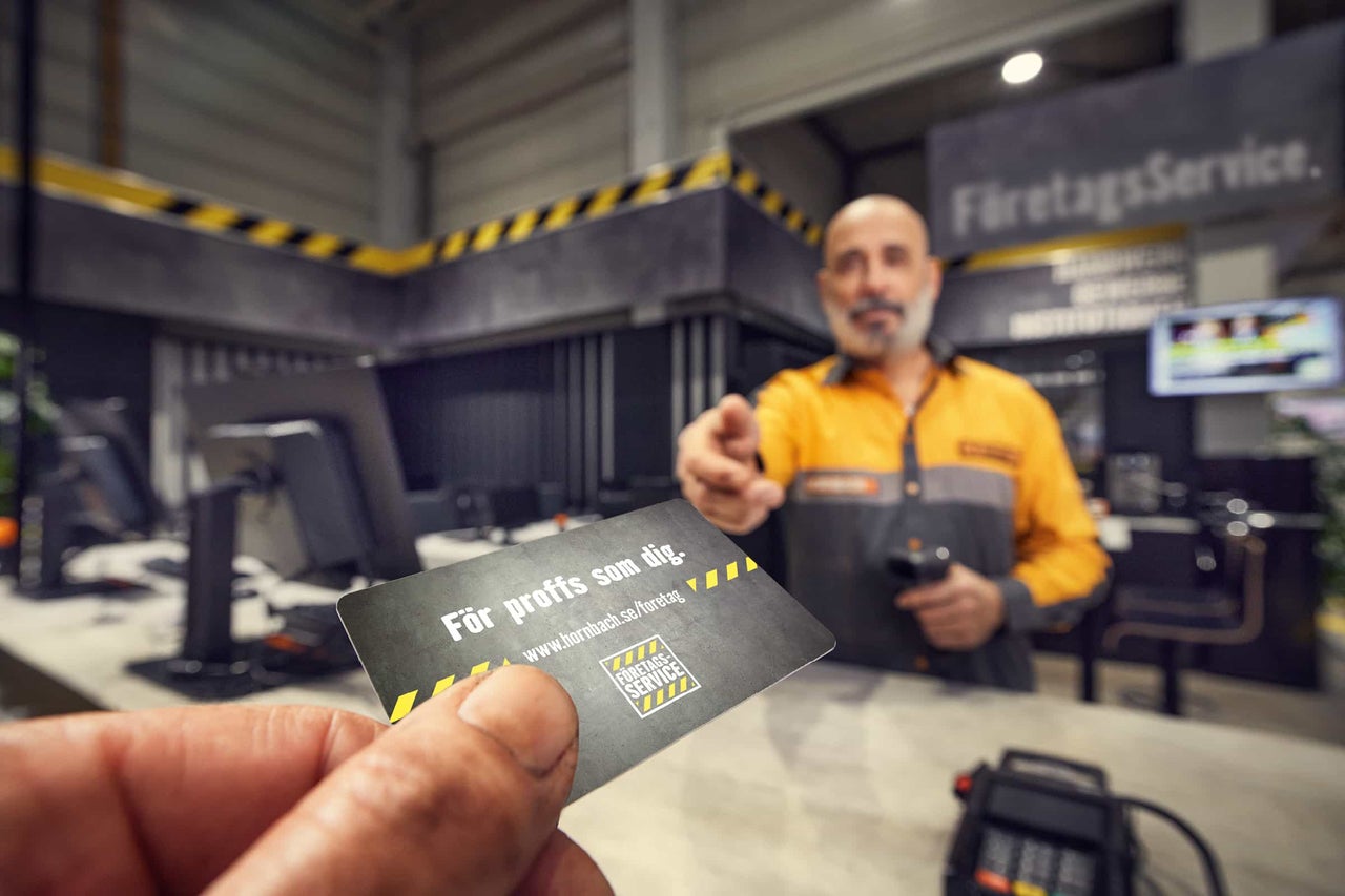 En hand håller ett Hornbach-kort för proffskunder framför en servicedisk för företagskunder med en medarbetare i bakgrunden.
