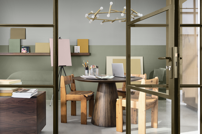 En stilren ateljé med ett runt träbord, trästolar, laptop och ett staffli framför en vägg med tvåfärgad design.