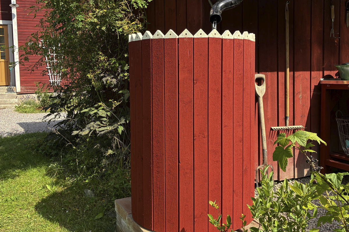 Regntunna av röda träribbor med dekorativa vita spetsar i en trädgård bredvid en trästuga och trädgårdsredskap.