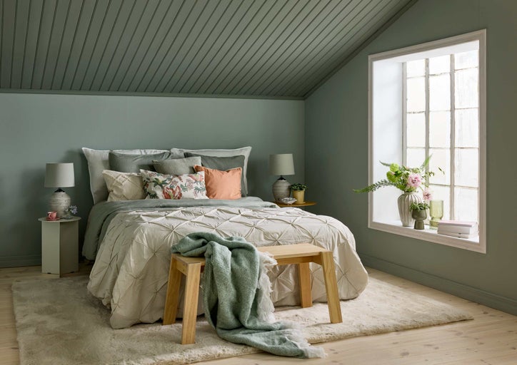 Mysigt sovrum med salviagröna väggar, panelat tak, säng med plisserat täcke, träbänk och mjuk matta på trägolv.