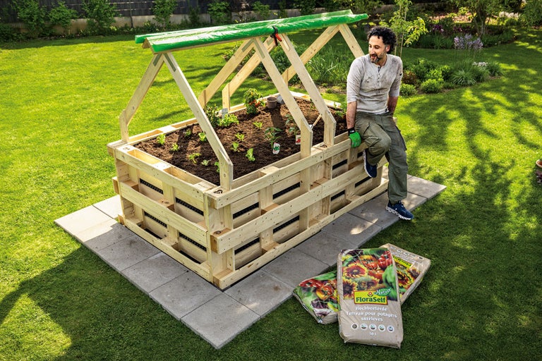 Upphöjd trädgårdssäng av trä med folietäckning och två säckar planteringsjord i trädgården.