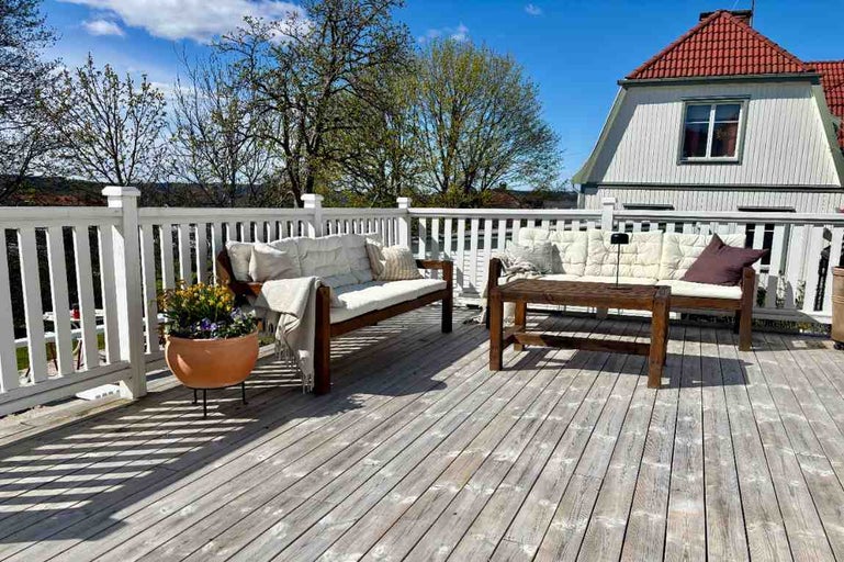Träterrass med vitt staket, trädgårdssoffor i trä med dynor, ett soffbord och en blomkruka.