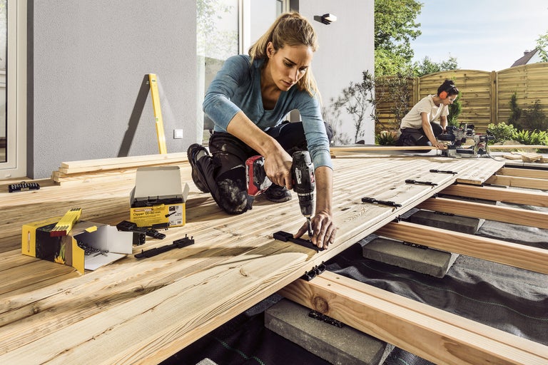 Byggande av en träterrass med trädgårdsplattor, underkonstruktion, fästklämmor och en person med en borrmaskin.